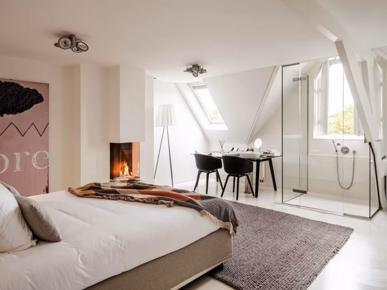 MANNA Signature Suite hotelkamer met een bed, een open haard, een ruime inloopdouche met regendouche en een wastafel met twee stoelen, onder een schuin dak met dakramen en een interieur in lichte tinten.