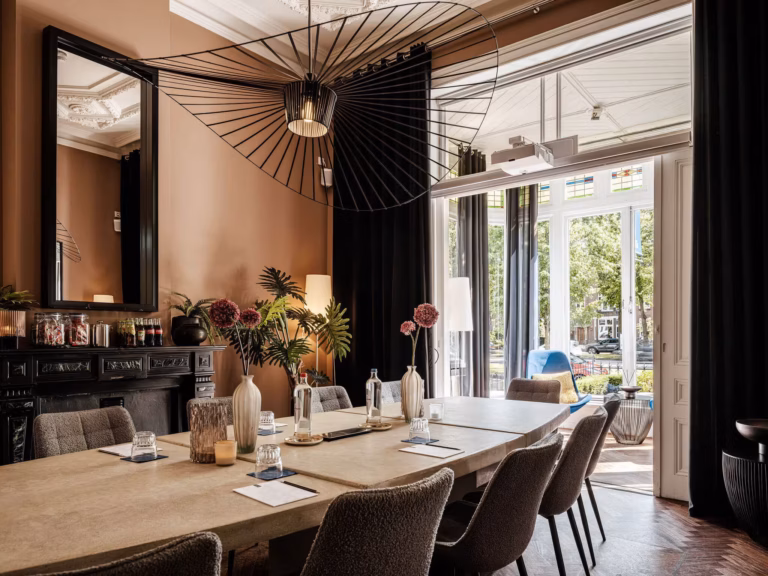 Blue Singel Boardroom met veel licht voor vergaderingen met een vergadertafel, gestoffeerde stoelen, een designhanglamp, een schouw, bloemen en glazen schuifdeuren naar een zithoek met glas-in-loodramen en uitzicht op Nijmegen.