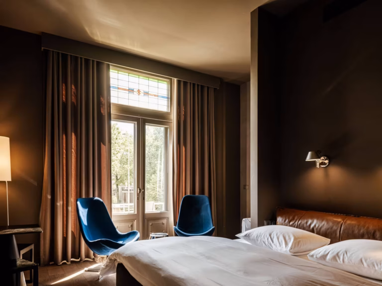 Casual Room van Boutique Hotel Blue met een tweepersoonsbed met leren hoofdbord, twee blauwe fauteuils, donkere muren en gordijnen en openslaande deuren met glas-in-loodraam en uitzicht op groen en Nijmegen.